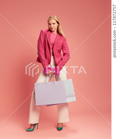 Full-length portrait of elegant young woman in stylish clothes standing with big shopping bag against pink studio background Full-length portrait of elegant young woman in stylish clothes standing with big shopping bag against pink studio background 117872757