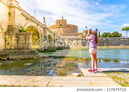 A young girl captures a photo of Castel SantAngelo and a bridge in Rome with her smartphone on a bright sunny day. Rome, Italy 117874716
