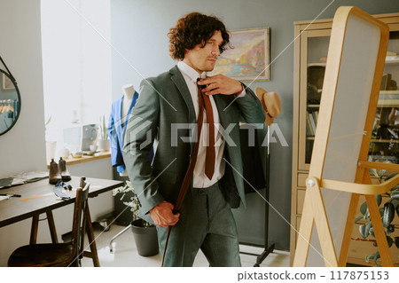 Curly haired male model dressed in gray suit and choosing tie in front of vertical mirror 117875153
