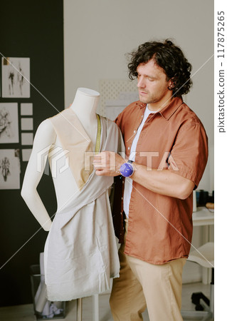 Caucasian curly haired tailor standing next to white plastic mannequin and measuring cotton fabric Caucasian curly haired tailor standing next to white plastic mannequin and measuring cotton fabric 117875265