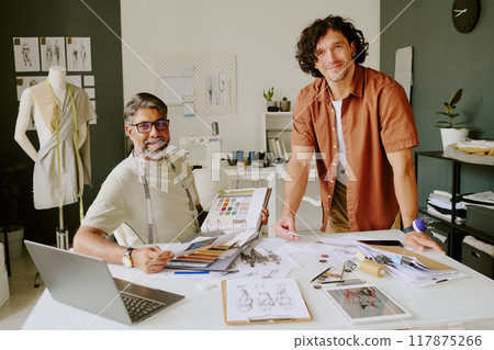 Portrait of mature designer with album in hands sitting at work desk and tailor standing next to desk while posing for photo Portrait of mature designer with album in hands sitting at work desk and tailor standing next to desk while posing for photo 117875266