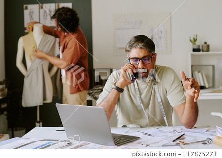 Indian tailor sitting at work desk and speaking on smartphone while his intern doing darts on mannequin 117875271
