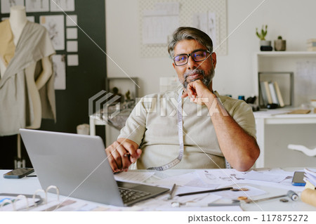 Famous Indian tailor sitting at white table and posing for portrait in own atelier Famous Indian tailor sitting at white table and posing for portrait in own atelier 117875272