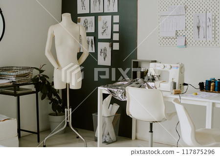 Creamy colored mannequin standing next to white table with metallic piece of fabric and white sewing machine Creamy colored mannequin standing next to white table with metallic piece of fabric and white sewing machine 117875296