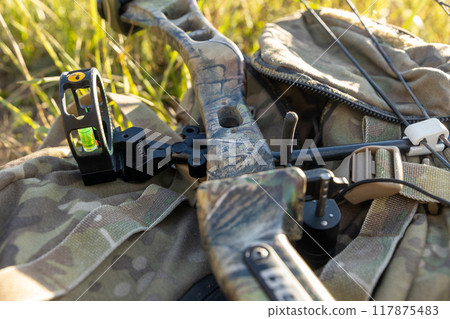 In a sunlit clearing, a camouflaged hunting compound bow rests on the backpack, poised for action 117875483