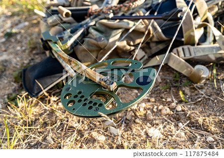 In a sunlit clearing, a camouflaged hunting compound bow rests on the send ground, poised for action 117875484