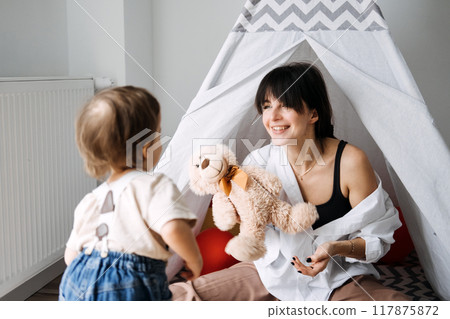 Mother playing with toddler using a toy bear in an indoor tent. Parent-child bonding, importance of play, relationship building, emotional connection, quality time 117875872