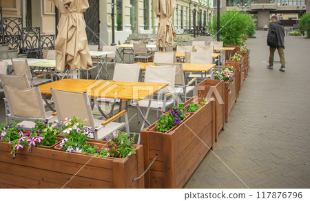 Early summer morning, outdoor outdoor cafe with wooden furniture and flower boxes and a lone passerby Early summer morning, outdoor outdoor cafe with wooden furniture and flower boxes and a lone passerby 117876796