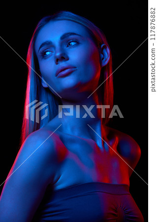 Young beautiful woman with flawless, clear skin showcasing impact of dedicated skincare routine against black studio background in neon light 117876882