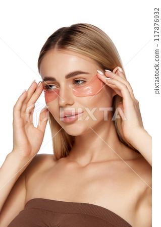 Close-up of young woman showing her flawless skin, using undereye patches, reflecting dedication to skincare and pursuit of natural, youthful beauty. 117876932