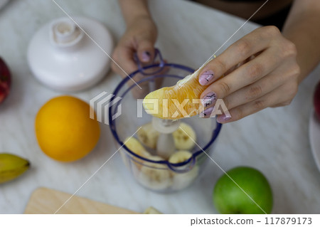 Orange slice in girl hands against background of fruits and blender on kitchen table at home, girl preparing sports cocktail with vitamins at home 117879173
