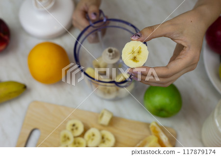 Piece of banana in hand of sporty girl against background of blender with fruits while preparing a sports cocktail. Healthy drink with vitamins from fruits and milk in blender at home kitchen Piece of banana in hand of sporty girl against background of blender with fruits while preparing a sports cocktail. Healthy drink with vitamins from fruits and milk in blender at home kitchen 117879174