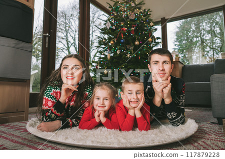 Smiling family portrait in festive sweaters laying under decorated Christmas tree at home during holiday. Happy parents and kids cuddling, feeling excited. Family time together. Happy childhood. Smiling family portrait in festive sweaters laying under decorated Christmas tree at home during holiday. Happy parents and kids cuddling, feeling excited. Family time together. Happy childhood. 117879228