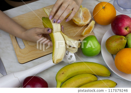 Female hands peeling banana on kitchen table with fruits on background. Girl preparing sports fruit cocktail. Healthy nutrition and lifestyle at home 117879246