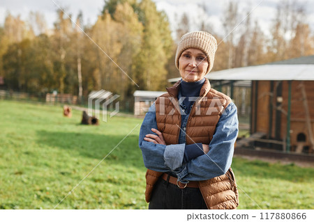 Waist up portrait of smiling mature female farmer in warm clothes with arms crossed looking at camera while posing at countryside farm during autumn season, copy space 117880866