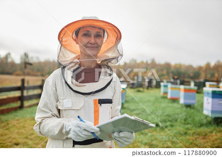 Medium portrait shot of smiling middle aged female beekeeper in protective bee suit holding notepad standing in meadow with hives against grey autumn sky at apiary farm, copy space 117880900