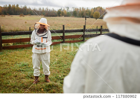 Full shot of middle aged female beekeeper in protective bee suit writing in notepad standing in meadow against autumn forest at apiary farm, copy space 117880902