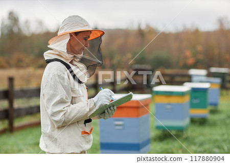 Side view of mature female beekeeper in protective bee suit writing in notepad while working around colorful wooden hives at autumn apiary farm, copy space Side view of mature female beekeeper in protective bee suit writing in notepad while working around colorful wooden hives at autumn apiary farm, copy space 117880904