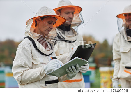 Side view of serious middle aged female apiarist in protective bee suit writing in notepad while keeping beehives with colleagues at apiary farm, copy space 117880906