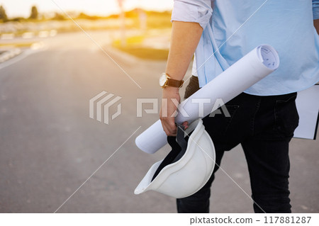 Unrecognisable engineer, architect or worker holds in hand protective white hard hat and blueprint on multi-apartment residential complex background, sunlighting. Concept of new real estate. 117881287