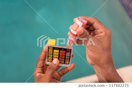 Testing the chlorine and PH of swimming pool. Close up of person testing the ph of the water with mini tester in swimming pool. Hand testing pool water for chlorine with PH mini tester 117882275