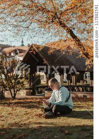Professional Photographer Taking Picture Of Beautiful Autumn Park. Man Professional Photographer Sit With Camera And With Smartphone In Autumn Park. Retouched, Vibrant Colors, Brownish Tones. Professional Photographer Taking Picture Of Beautiful Autumn Park. Man Professional Photographer Sit With Camera And With Smartphone In Autumn Park. Retouched, Vibrant Colors, Brownish Tones. 117882591