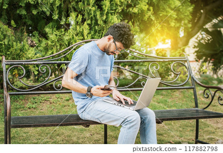 Young man in a park with laptop and cell phone. Freelancer man sitting in a park using laptop and cellphone. Man in a park working online with laptop. Relaxed man working with laptop outdoor 117882798