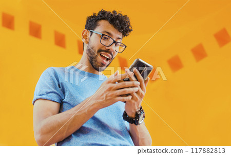 Happy guy sending a message with a cell phone near a yellow wall outdoors. Lifestyle of handsome bearded man using cell phone outdoors, Low angle view of young man using cell phone outdoors Happy guy sending a message with a cell phone near a yellow wall outdoors. Lifestyle of handsome bearded man using cell phone outdoors, Low angle view of young man using cell phone outdoors 117882813
