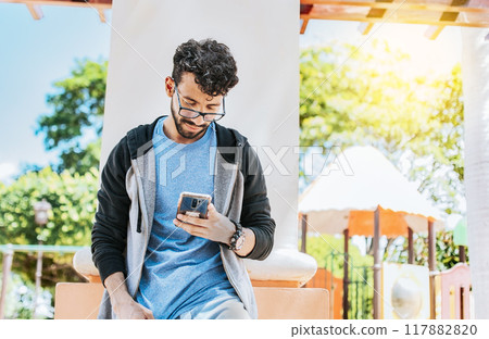 Smiling handsome man using a phone leaning on a wall in a park, Handsome guy in glasses leaning on a wall chatting on his cell phone, Teenage male leaning on a wall sending a text message Smiling handsome man using a phone leaning on a wall in a park, Handsome guy in glasses leaning on a wall chatting on his cell phone, Teenage male leaning on a wall sending a text message 117882820