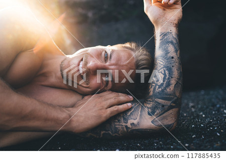Cheerful tattooed man lying on sandy coast Cheerful tattooed man lying on sandy coast 117885435