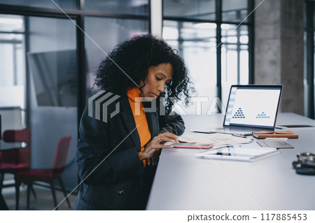Focused Black businesswoman in her 30s, with curly hair, reviews documents at her office desk 117885453