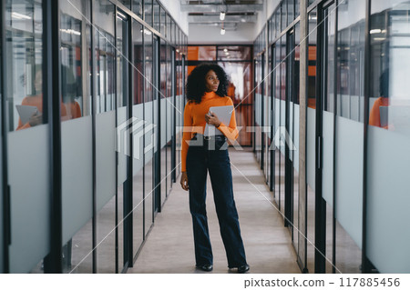 Black businesswoman in orange top and black trousers walking with camera in modern office hallway Black businesswoman in orange top and black trousers walking with camera in modern office hallway 117885456