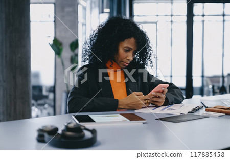 Diligent Black businesswoman in 30s, wearing gray blazer and orange turtleneck, engaged with smartphone Diligent Black businesswoman in 30s, wearing gray blazer and orange turtleneck, engaged with smartphone 117885458