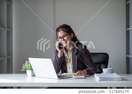 Beautiful accountant talking on the phone and calculating financial figures and calculating taxes for the company. Beautiful accountant talking on the phone and calculating financial figures and calculating taxes for the company. 117889174