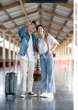 Happy Asian couple holding suitcases and camera preparing to wait for train at train station for vacation trip together. 117889226