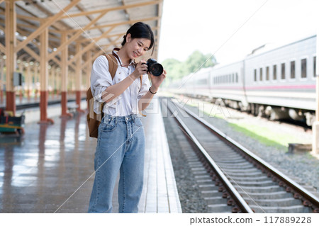 Young Asian woman holds a camera and takes photos of a train while waiting for the train at the train station to travel. 117889228
