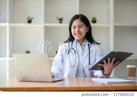young asian lady doctor in white medical uniform with stethoscope using computer laptop talking video conference call with patient at desk in health clinic or hospital. 117889229