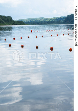 Orange buoys floating on a calm lake with forested shore in the background. Orange buoys floating on a calm lake with forested shore in the background. 117890170