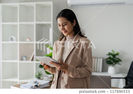 young asian woman, company worker in suit, smiling and holding digital tablet, standing over working space background 117890333