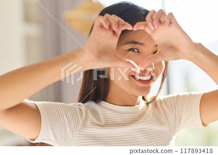 Smiling Woman Framing Her Eye With Heart-Shaped Hands In Bright Room Smiling Woman Framing Her Eye With Heart-Shaped Hands In Bright Room 117893418