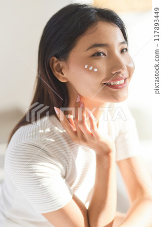 Face Cream. Woman Applying Skincare Cream with Radiant Smile in Sunlit Room 117893449