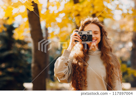 Smiling young woman with the photo camera shooting the forest trees with the yellow leaves at sunset Smiling young woman with the photo camera shooting the forest trees with the yellow leaves at sunset 117897004