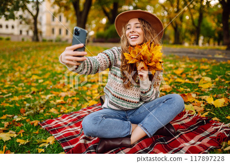 Selfie time. Beautiful woman takes selfie on smartphone in hat on meadow in autumn park on plaid. Selfie time. Beautiful woman takes selfie on smartphone in hat on meadow in autumn park on plaid. 117897228