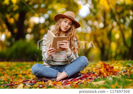 Beautiful curly woman in hat on mat with book in autumn park. Relaxation, solitude with nature. Beautiful curly woman in hat on mat with book in autumn park. Relaxation, solitude with nature. 117897308