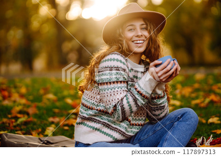 Curly woman with thermos in clearing among fallen leaves in autumn park, enjoying hot drink. 117897313
