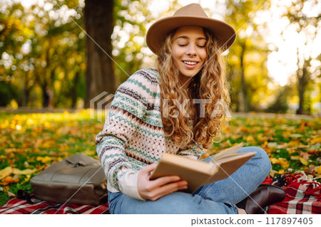 Beautiful curly woman in hat on mat with book in autumn park. Relaxation, solitude with nature. 117897405