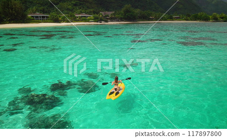 Paddling a yellow kayak in turquoise waters towards a tropical resort, a woman enjoys nature's beauty, blending adventure and relaxation in a serene lagoon Paddling a yellow kayak in turquoise waters towards a tropical resort, a woman enjoys nature's beauty, blending adventure and relaxation in a serene lagoon 117897800