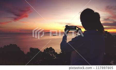 Woman silhouette stand on hill make photo colorful evening seascape scene. Person enjoy dramatic beautiful ocean sunset sky. Travel to Thailand, summer vacation. Lifestyle holiday concept. Back view Woman silhouette stand on hill make photo colorful evening seascape scene. Person enjoy dramatic beautiful ocean sunset sky. Travel to Thailand, summer vacation. Lifestyle holiday concept. Back view 117897805