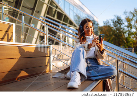 Young woman in headphones with a laptop on a city street. ?nline training. Freelancer. 117898687