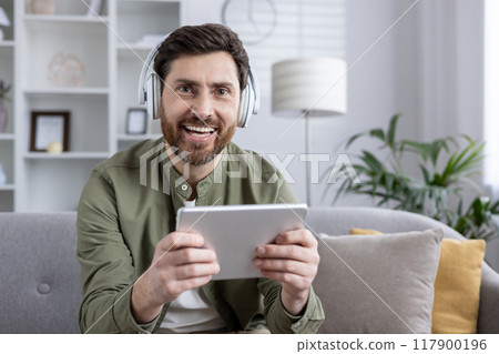 Smiling man wearing headphones holding tablet while sitting on sofa in brightly lit living room. Person enjoying digital content, technology, and relaxation during leisure time. Smiling man wearing headphones holding tablet while sitting on sofa in brightly lit living room. Person enjoying digital content, technology, and relaxation during leisure time. 117900196
